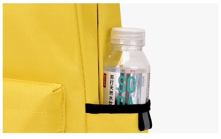 Side detail of the yellow backpack: a black elastic retention band grips a plastic beverage bottle prop against the panel, finished with a small grey webbing tab