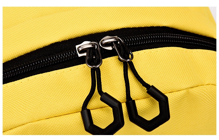 Macro detail of a yellow school backpack's double-zip top: two metal sliders side by side on black tape, each threaded with a black cord loop ending in a moulded geometric pull tab