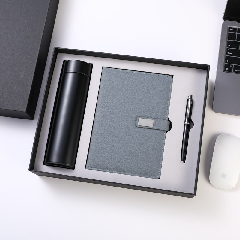 Black-and-grey variant of the boxed trio: matte black tumbler, slate grained journal with blank metal plate, black-and-chrome pen, open rigid box with lid set aside on a white desk with laptop and mouse props