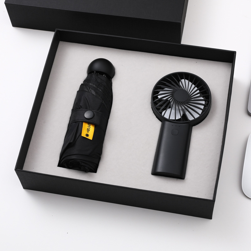 Black folding umbrella with snap strap and black handheld fan seated in cut-outs of an open matte black gift box