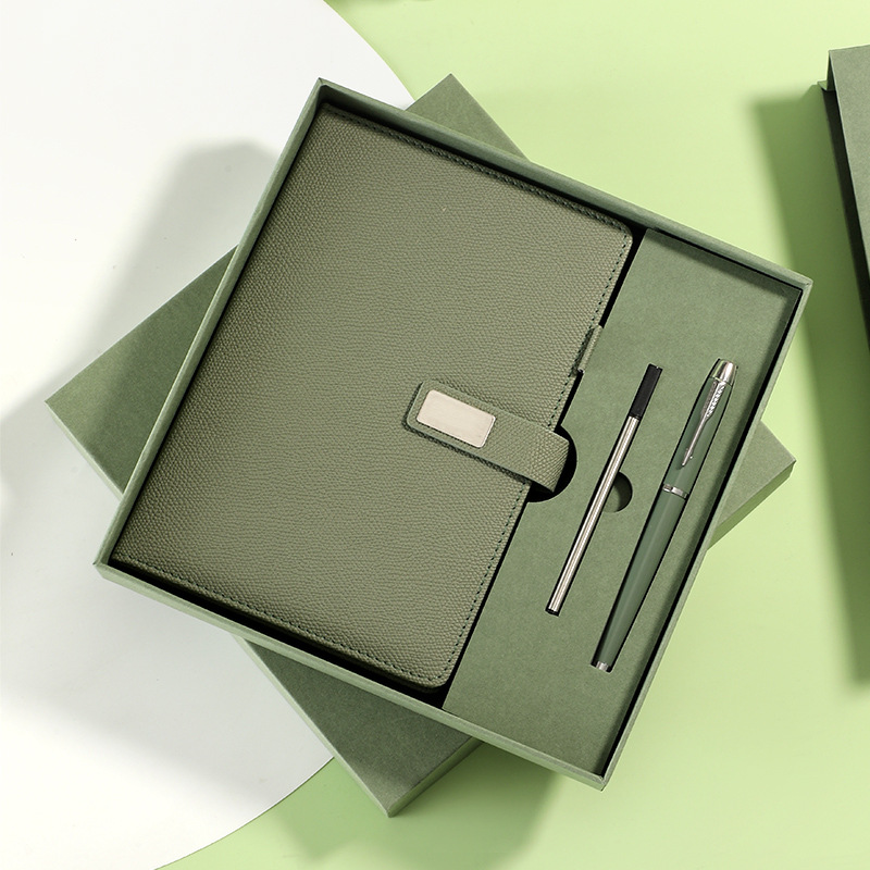 Sage green journal-and-pen gift set in an open rigid box with fitted tray, photographed at an angle on a white disc over a mint ground; textured cover, blank metal strap plate, green pen with chrome clip, slim silver refill rod