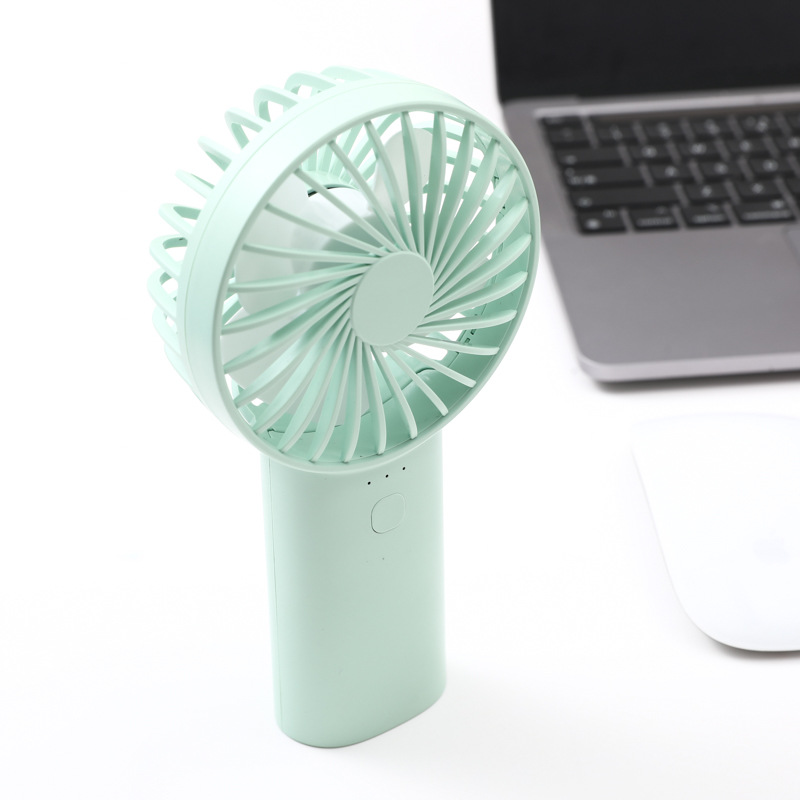 Mint green handheld fan standing on its handle, spiral safety grille over white blades, single button and three indicator dots on a desk