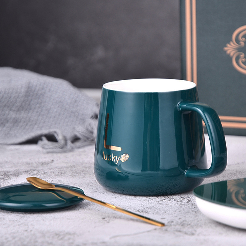 Teal glazed mug with gold monogram sample print and white interior, gold spoon on a small teal saucer, patterned dish and foil-trimmed green gift box panel behind, styled on a pale stone surface with a draped grey textile