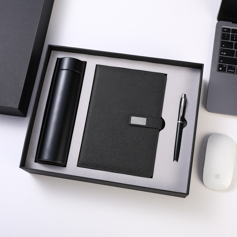 Black boxed trio flat-lay on white: matte vacuum tumbler, crosshatch journal with blank silver plate on its closure strap, chrome-trim pen in a light-grey fitted insert, beside a cropped laptop and mouse styling the desk scene