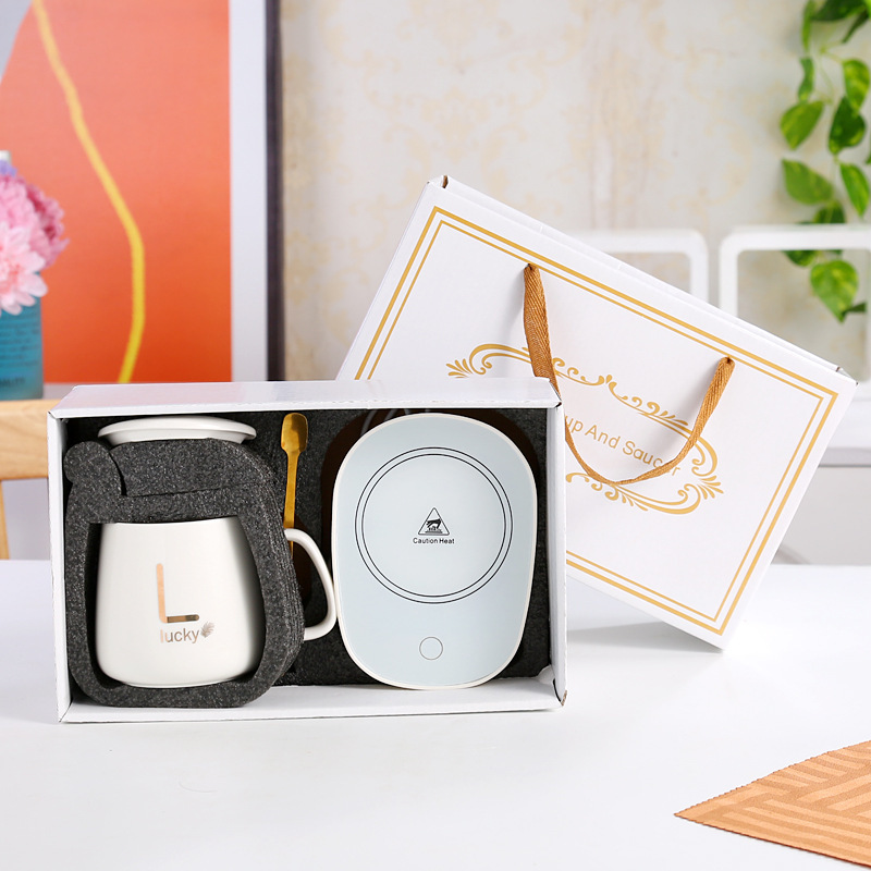Black lidded mug with gold monogram sample print, gold spoon and black warming coaster printed with a centred heat warning in white, boxed in a dark green gift box with grey foam cut-outs, gold-trimmed ribbon-handle bag behind