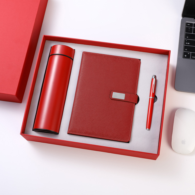 Red boxed trio on white: satin red flask, red grained notebook with blank engraving plate and red pen with polished trim in a fitted insert, lid stood aside, laptop and mouse as desk props