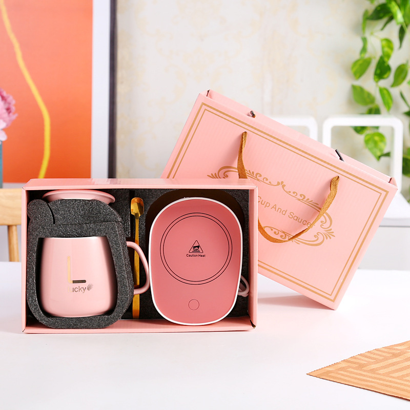 Pink edition of the same box set: glossy pink lidded mug with gold monogram sample, gold spoon in a foam slot, pink warming coaster with white heat caution marking and white cord, matching pink bag with gold scrollwork and rope handles