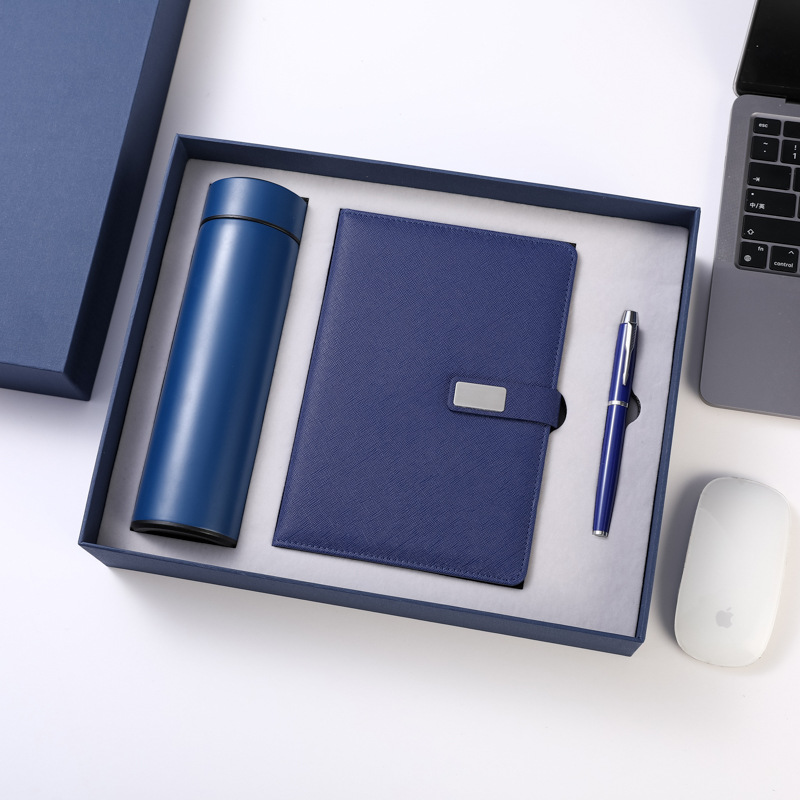 Navy boxed trio on white: matte navy tumbler, cobalt grained journal with blank silver plate, navy-barrel pen with chrome clip in the grey insert, lid at corner, cropped laptop keyboard and mouse staging