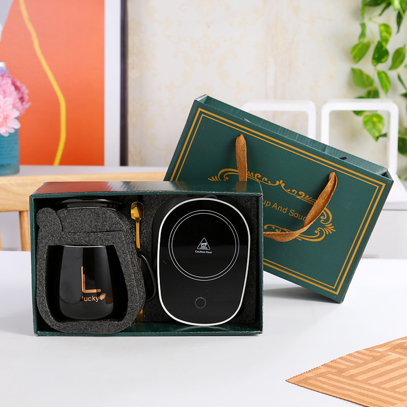 Teal glazed lidded mug with gold monogram sample print and gold leaf glyph, gold spoon resting on a matching teal saucer, second gold-patterned dish at frame edge, foil-trimmed dark green gift box standing behind on a grey stone surface