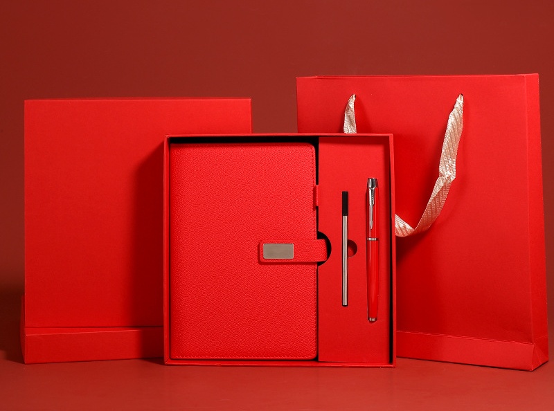 Deep red gift-set staging on a crimson backdrop: upright rigid lid, open tray box holding a red leather-grain journal with blank metal plate and two pen components, and a red ribbon-handle carrier bag