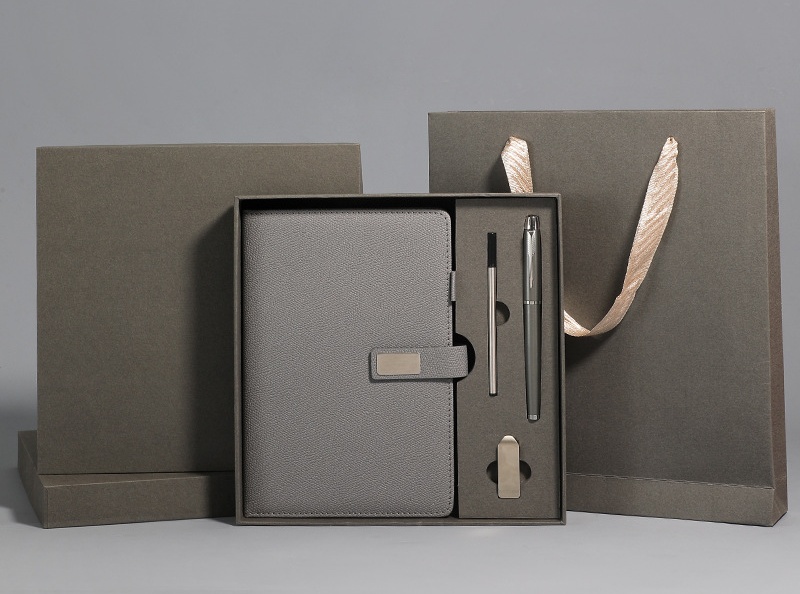 Warm grey executive set: closed rigid box with lid stood behind, open tray with grained notebook, blank metal plate, gunmetal pen, black stylus and brushed metal USB bar, plus a matching ribbon-handle bag at right